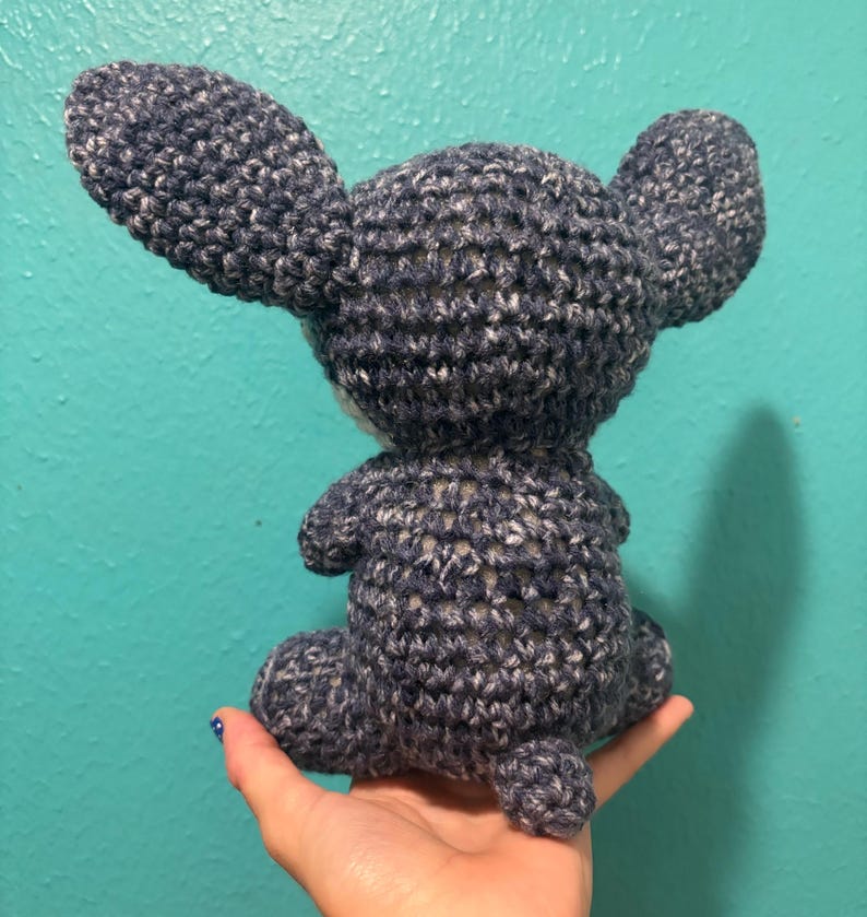 Puede incluir: Un peluche de ganchillo hecho a mano, visto desde atr&aacute;s, en posici&oacute;n sentada. El juguete es de color azul oscuro con motas blancas, con orejas grandes y una cola peque&ntilde;a. El juguete se sostiene en una mano.