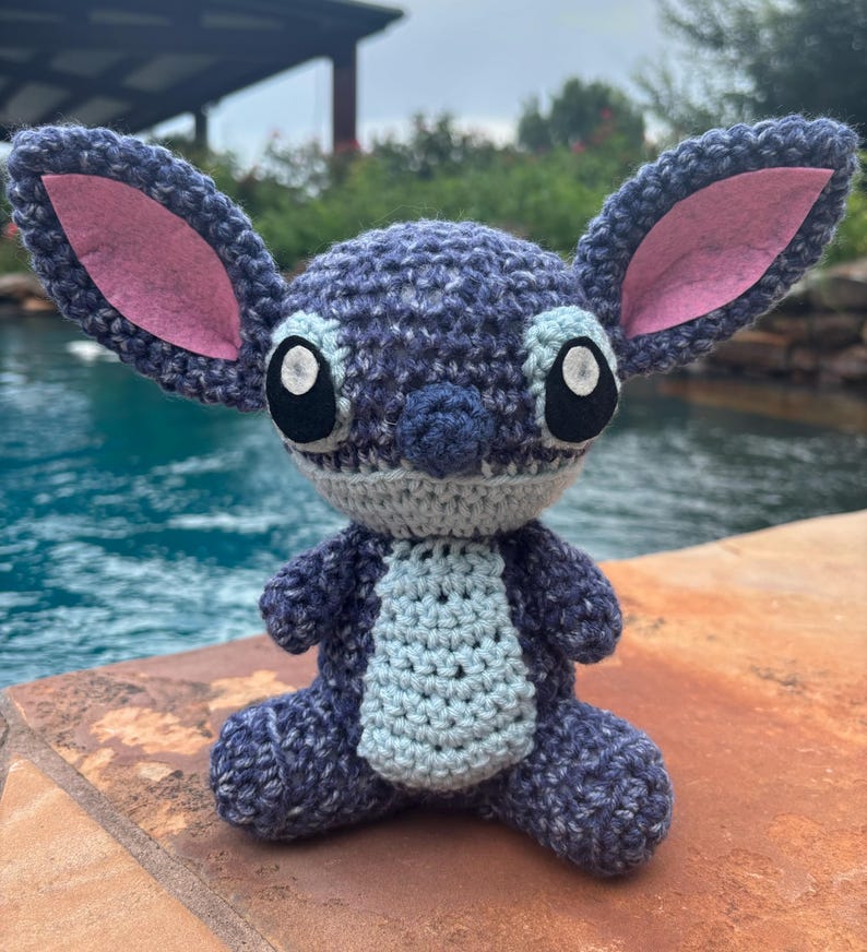 Puede incluir: Un peluche hecho a mano con ganchillo en forma de un extraterrestre azul con orejas grandes y ojos grandes. El juguete tiene orejas internas rosas, una barriga azul claro y est&aacute; sentado en una superficie marr&oacute;n cerca de una piscina.