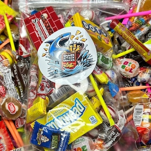 Könnte beinhalten: Eine durchsichtige Plastiktüte gefüllt mit einer Vielzahl von bunten Süßigkeiten, darunter Lutscher, Gummibärchen und harte Bonbons. Die Tüte enthält außerdem mehrere bunte Plastikstäbchen und einen kleinen weißen Aufkleber mit dem Text "SHARK ATTACK SNACKS".