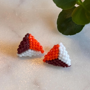 May include: A pair of handmade beaded triangle earrings. Each earring has a geometric design with white, orange, and burgundy beads. The earrings are displayed on a white marble surface.