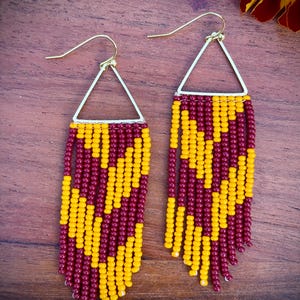 May include: A pair of beaded earrings. Each earring has a gold-coloured triangular top and a fringe of maroon and yellow beads. The earrings are displayed on a wooden surface.