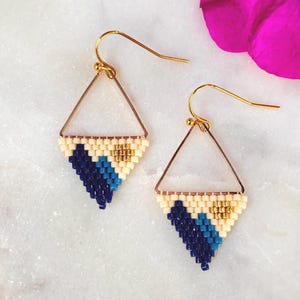 Puede incluir: Un par de pendientes triangulares con marco de metal dorado y un patrón de cuentas tejidas. Las cuentas están dispuestas en un diseño geométrico con colores crema, azul verdoso y azul oscuro, acentuado por un pequeño grupo de cuentas doradas.