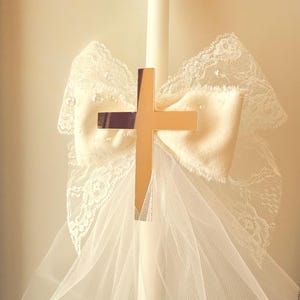 Könnte beinhalten: Eine weiße Kerze, verziert mit einem goldenen Kreuz, einer cremefarbenen Schleife und zarter Spitze und Tüll. Das Kreuz ist mittig auf der Kerze platziert, wobei die Schleife und die Spitze eine dekorative Note hinzufügen. Der Tüll fällt herab und erzeugt einen weichen, eleganten Look.