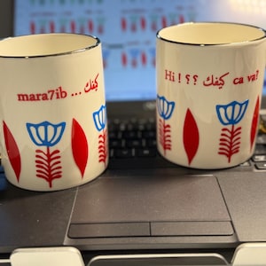 Puede incluir: Dos tazas de cerámica blanca con ribete dorado y diseño floral. Una taza dice "Hi!  ?  كيفك ca va?" y la otra taza dice "كيفك... mara7ib".