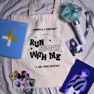 Könnte beinhalten: Eine weiße Tragetasche mit dem Text "TOMORROW X TOGETHER" und "RUN AWAY WITH ME 9 AND THREE QUARTERS" darauf gedruckt. Die Tasche ist gefüllt mit verschiedenen Gegenständen, darunter ein blaues und weißes Album, ein hellblauer und weißer Lichtschläger und mehrere kleine Foto-Karten.