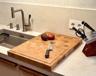 May include: A wooden cutting board with a checkered pattern, a knife, two avocados, and a red tomato. The board is on a white countertop next to a stainless steel sink and a knife block.