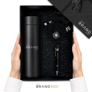 K&ouml;nnte beinhalten: Eine schwarze Geschenkbox mit einer mattschwarzen zylindrischen Trinkflasche mit dem Text YOUR BRAND HERE, einem schwarzen Kugelschreiber mit dem Text YOUR LOGO HERE und einem schwarzen Multi-Port-Ladekabel mit einem runden Anh&auml;nger YOUR LOGO HERE.