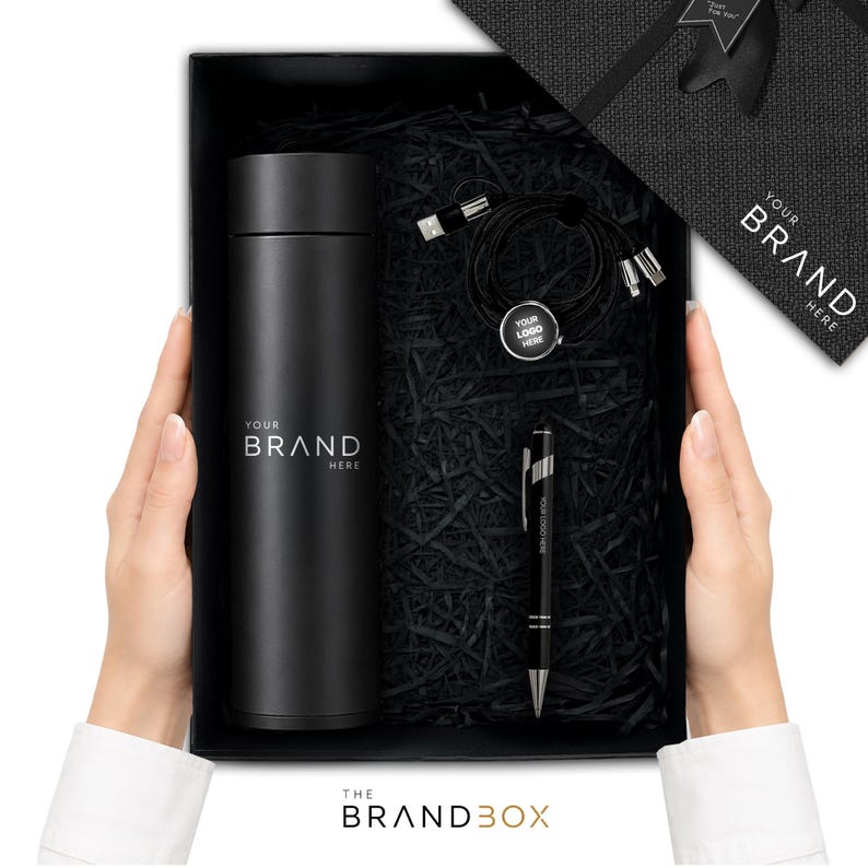 K&ouml;nnte beinhalten: Eine schwarze Geschenkbox mit einer mattschwarzen zylindrischen Trinkflasche mit dem Text YOUR BRAND HERE, einem schwarzen Kugelschreiber mit dem Text YOUR LOGO HERE und einem schwarzen Multi-Port-Ladekabel mit einem runden Anh&auml;nger YOUR LOGO HERE.