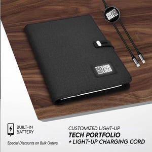 May include: Black tech portfolio with a light-up logo and a light-up charging cord. The portfolio has a built-in battery and a clasp closure. The text "YOUR LOGO" and "HERE" are visible on the portfolio and charging cord.