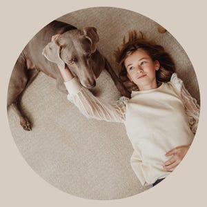 May include: A young person with long blonde hair is lying on a light brown carpet with a gray Weimaraner dog. The person is wearing a white sweater with sheer sleeves and is looking at the camera. The dog is looking at the person.