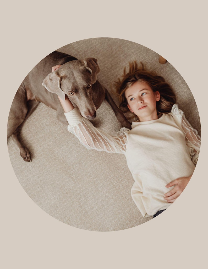 May include: A young person with long blonde hair is lying on a light brown carpet with a gray Weimaraner dog. The person is wearing a white sweater with sheer sleeves and is looking at the camera. The dog is looking at the person.