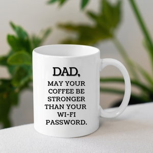 May include: White ceramic coffee mug with the text "DAD, MAY YOUR COFFEE BE STRONGER THAN YOUR WI-FI PASSWORD." The mug has a curved handle and is set against a blurred background of green foliage.