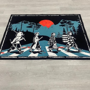 Puede incluir: Alfombra rectangular negra con una parodia de la portada del &aacute;lbum Abbey Road con personajes de la serie de anime Demon Slayer cruzando un paso de cebra. El fondo incluye un sol rojo, monta&ntilde;as y &aacute;rboles. La alfombra tiene un borde negro.