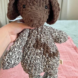 May include: A handcrafted crochet dog toy. The dog's head and ears are brown, with a speckled white and brown body. The toy is made from soft yarn, suitable for a child's play or as a decorative item.
