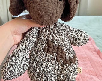 May include: A brown and white crocheted dog plush toy. The dog has a brown head and ears, and a white body with brown accents. The toy is sitting on a pink surface.