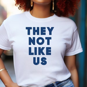 May include: A white t-shirt with the text "THEY NOT LIKE US" printed in blue. The model is wearing the shirt and has red and orange curly hair, gold-rimmed sunglasses, and gold earrings.