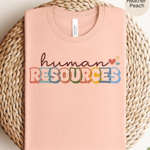 Pode incluir: Uma camiseta cor pêssego com o texto "human resources" em letras coloridas de estilo retrô. O texto está em uma fonte divertida e caprichosa com um coração e pequenos corações acima da palavra "human".