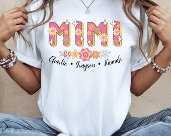 May include: White t-shirt with the word "MIMI" in pink letters with floral accents and the names "Charlie, Grayson, Hannah" underneath.