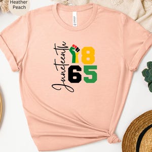 Puede incluir: Una camiseta de color melocot&oacute;n jaspeado con el texto "Juneteenth 1865" y un gr&aacute;fico de un pu&ntilde;o levantado en negro, rojo, verde y amarillo.