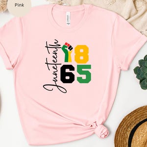 Puede incluir: Camiseta rosa con el texto "Juneteenth 1865" y un pu&ntilde;o levantado en rojo, negro y verde.