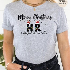 Pode incluir: Uma camiseta cinza com o texto "Merry Christmas HR approved" em preto. As letras "HR" são decoradas com luzes de Natal e chapéus de Papai Noel.