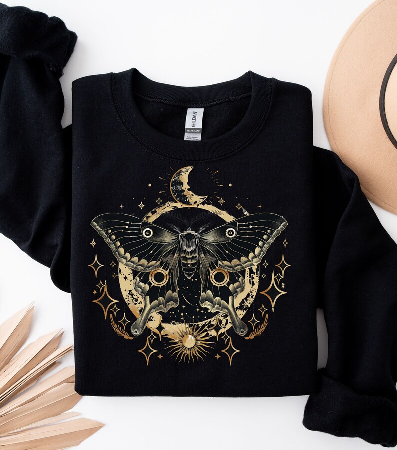 Pu&ograve; includere: Felpa nera con una grafica dorata di una falena, luna e stelle. La falena ha ali grandi ed &egrave; circondata da un cerchio di stelle e una luna crescente.
