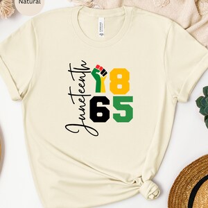 Puede incluir: Una camiseta de color natural con el texto "Juneteenth 1865" en letras negras y verdes. Un pu&ntilde;o negro con acentos rojos y amarillos est&aacute; en el centro del dise&ntilde;o.