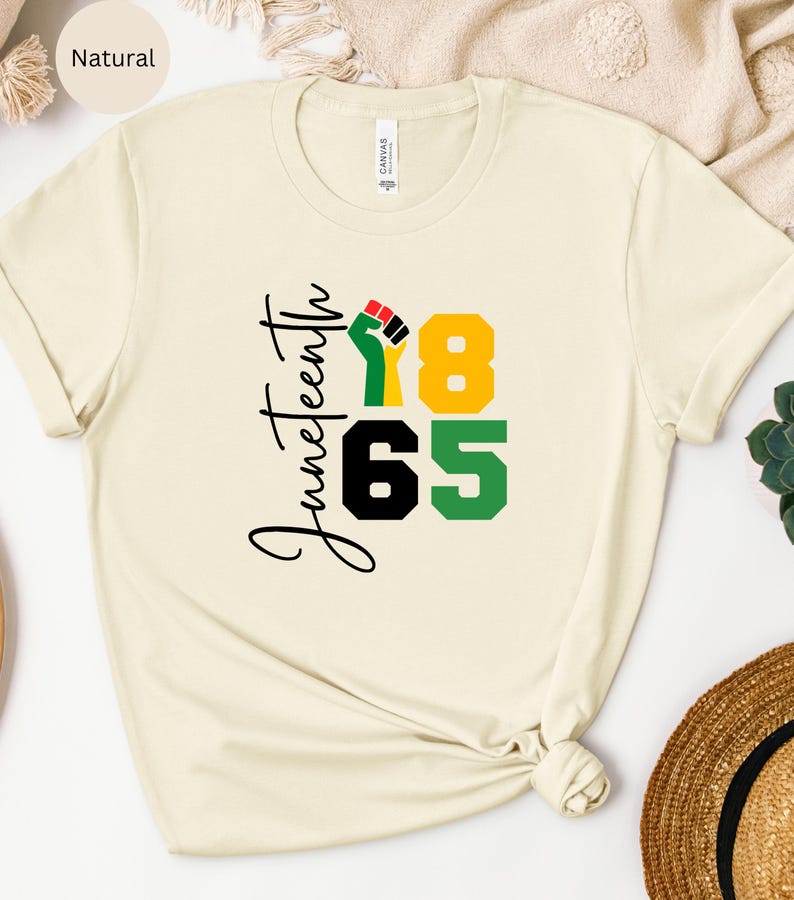Puede incluir: Una camiseta de color natural con el texto "Juneteenth 1865" en letras negras y verdes. Un pu&ntilde;o negro con acentos rojos y amarillos est&aacute; en el centro del dise&ntilde;o.