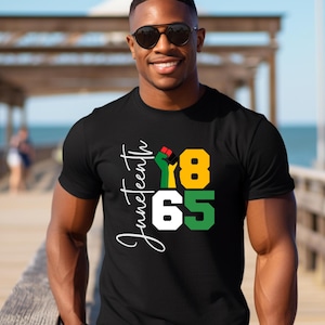 Puede incluir: Camiseta negra con un dise&ntilde;o gr&aacute;fico blanco que dice "Juneteenth 1865" con un pu&ntilde;o en alto en los colores de la bandera pan-africana.