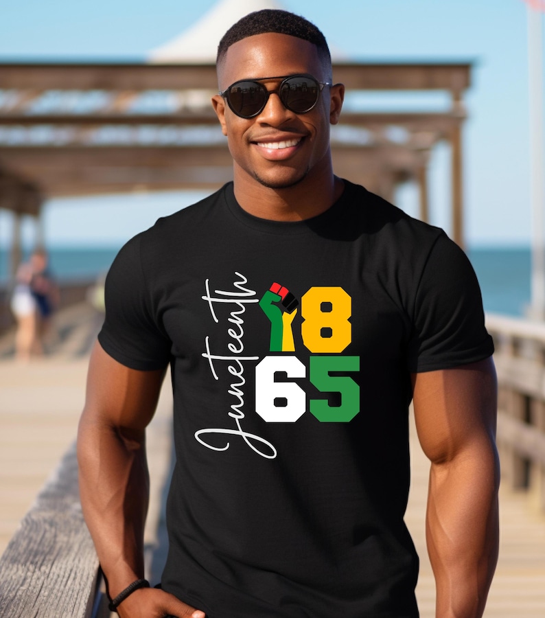 Puede incluir: Camiseta negra con un dise&ntilde;o gr&aacute;fico blanco que dice "Juneteenth 1865" con un pu&ntilde;o en alto en los colores de la bandera pan-africana.