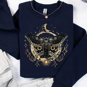 Pu&ograve; includere: Una felpa blu navy con una grafica dorata di una falena, una luna e stelle. La falena ha grandi ali nere con accenti dorati. La luna &egrave; a forma di mezzaluna e ha un contorno dorato. Le stelle sono piccole e dorate.