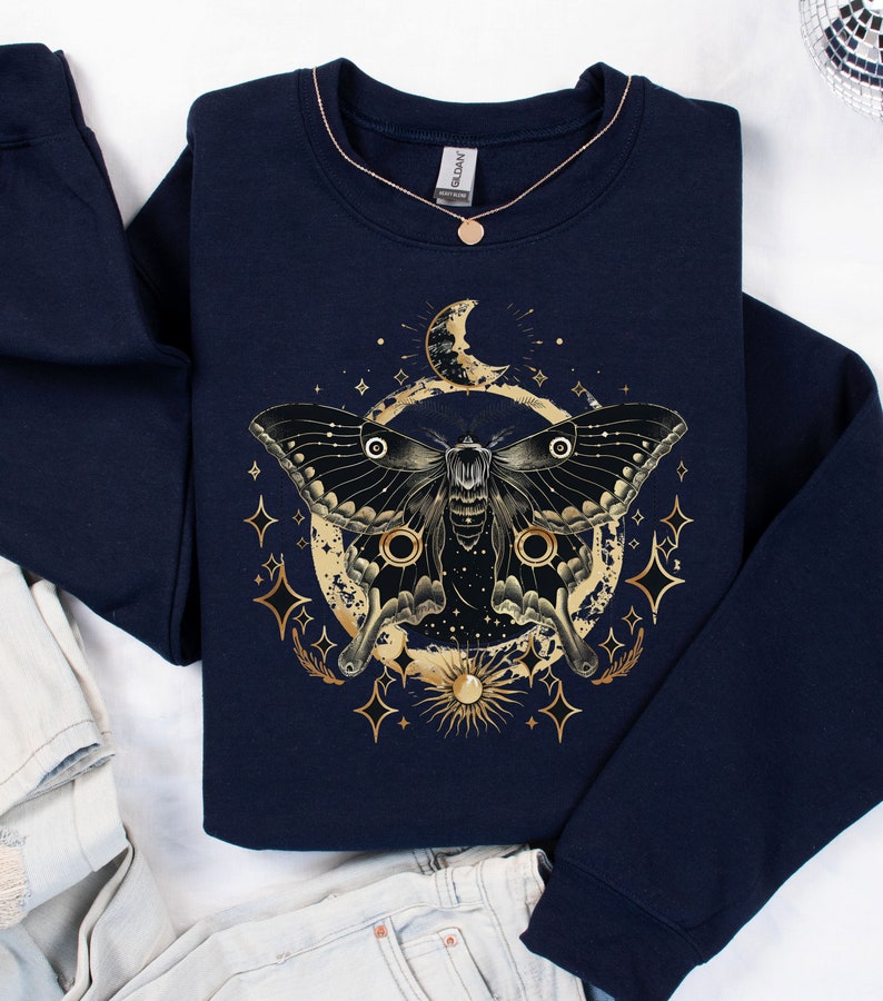 Pu&ograve; includere: Una felpa blu navy con una grafica dorata di una falena, una luna e stelle. La falena ha grandi ali nere con accenti dorati. La luna &egrave; a forma di mezzaluna e ha un contorno dorato. Le stelle sono piccole e dorate.