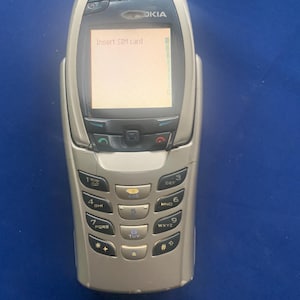 May include: A silver Nokia mobile phone with a rectangular screen displaying the text Insert SIM card. The device features a standard numeric keypad with twelve buttons, a central navigation control, and call answer and end buttons below the screen.