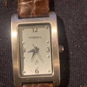 May include: A rectangular Fossil wristwatch with a silver-toned face, featuring the numbers 3, 6, 9, and 12, along with the Fossil logo and f2 text. The watch has a brown textured leather strap and a metallic silver-toned rectangular case.