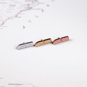 May include: Three metal train-shaped stud earrings in silver, gold, and rose gold. The earrings are small and detailed, with a rectangular body and a curved front. They are placed on a white surface with a map in the background.