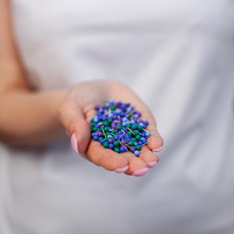 Puede incluir: Un pu&ntilde;ado de chinchetas de colores en tonos azules, verdes y morados. Las chinchetas tienen cabezas peque&ntilde;as y redondas y v&aacute;stagos plateados. Las chinchetas se sostienen en una mano abierta sobre un fondo blanco.