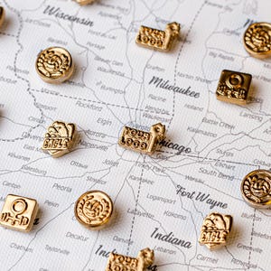 May include: A collection of small gold-colored metal pins featuring various embossed designs, including circular laurel wreaths, square symbols with a circle and bar, and rectangular shapes with the text "YOUR LOGO" or "EAGLE".