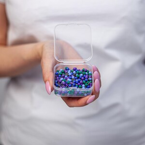 Puede incluir: Una peque&ntilde;a caja de pl&aacute;stico transparente llena de chinchetas de colores. Las chinchetas tienen cabezas redondas en tonos de azul, morado y verde. La caja se sostiene abierta, mostrando el contenido. El fondo es blanco liso.