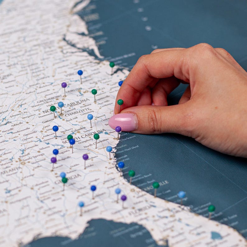 Puede incluir: Un mapa detallado del sureste de los Estados Unidos con numerosas chinchetas de colores que marcan ubicaciones. Una mano est&aacute; colocando una chincheta verde en el mapa. El mapa tiene un fondo azul-verdoso y es un producto con tem&aacute;tica de viajes.