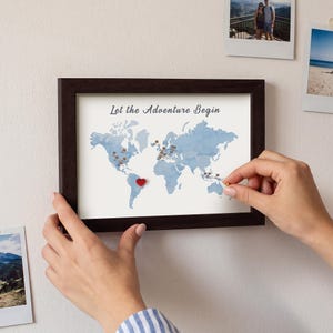May include: A framed world map print with a dark brown frame, featuring the text Let the Adventure Begin in gray script. The map has light blue continents on a white background, marked with small pins and a single red heart shape.