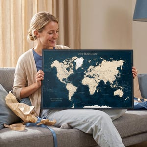 Può includere: Una mappa del mondo rettangolare incorniciata con uno sfondo blu scuro e masse terrestri color crema. Il testo "OUR TRAVEL MAP" è stampato in alto e "LIFE IS A JOURNEY BEST TRAVELED TOGETHER" è stampato nell'angolo in basso a sinistra.