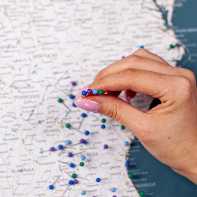 Puede incluir: Primer plano de un mapa con chinchetas. Una mano sostiene una fila de chinchetas de colores: moradas, azules y verdes. El mapa es blanco con texto y l&iacute;neas grises, sobre un fondo azul oscuro.