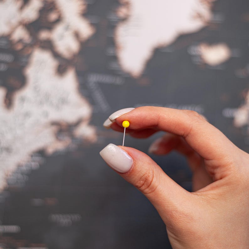 Puede incluir: Un chincheta amarilla sostenida entre los dedos, contra un fondo borroso de mapa del mundo. La chincheta tiene una cabeza peque&ntilde;a y redonda y un fino v&aacute;stago plateado. El mapa es en tonos grises y marrones.