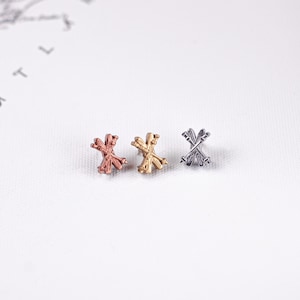 Puede incluir: Tres pequeños objetos decorativos de metal en oro rosa, oro y plata. Cada objeto tiene un diseño de esquís cruzados. Los objetos se muestran sobre una superficie blanca y texturizada. Los objetos son pequeños y detallados.