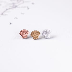 Può includere: Tre orecchini a lobo a forma di conchiglia in oro rosa, oro e argento. Gli orecchini sono piccoli, con un design dettagliato a conchiglia. Sono disposti in fila su uno sfondo bianco, suggerendo un tema di gioielli o accessori.