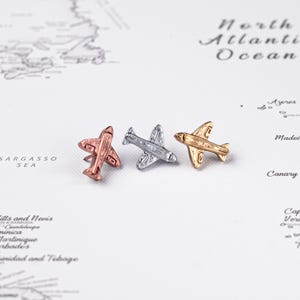 Könnte beinhalten: Drei kleine Flugzeug-Anstecknadeln in Roségold, Silber und Gold. Die Anstecknadeln befinden sich auf einer Karte des Nordatlantiks und des Karibischen Meeres. Jede Nadel hat Flügel, einen Rumpf und ein Heck.