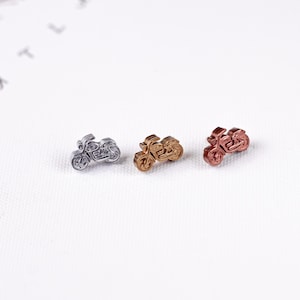 May include: Three miniature motorcycle charms in silver, gold, and copper colors. The charms are detailed with wheels, seats, and handlebars, and are arranged on a white surface. These charms could be used for jewelry making or other craft projects.