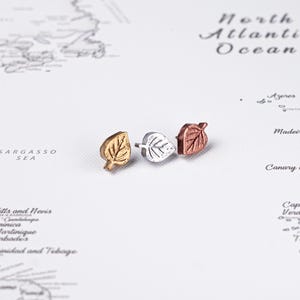 May include: Three leaf-shaped stud earrings in gold, silver, and copper tones. The earrings are arranged on a map background, showcasing their delicate design and metallic finishes. These earrings are a nature-inspired accessory.