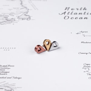 May include: Three metal map pin push pins in rose gold, gold, and silver. The pins are shaped like teardrops with a heart-shaped cutout. The pins are on a white map background with the words "North Atlantic Ocean" visible.