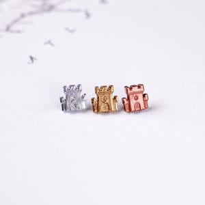 May include: Three miniature castle-shaped stud earrings in silver, gold, and rose gold. The earrings feature turrets and arched doorways, set against a white background. These small, decorative earrings are suitable for everyday wear.